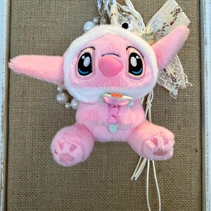 May include: A pink plush toy of a character with large black eyes, wearing a white hooded outfit. The toy has large ears and is attached to a metal heart-shaped clip with a pearl and ribbon accent. The background is a burlap texture.