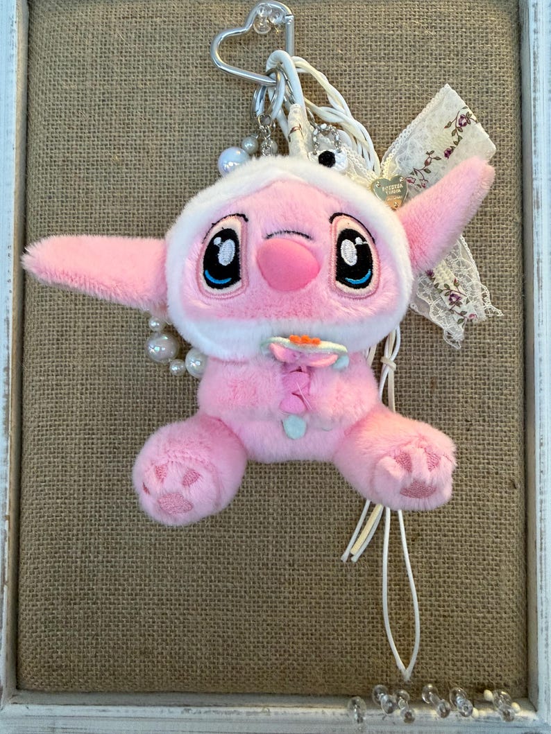 May include: A pink plush toy of a character with large black eyes, wearing a white hooded outfit. The toy has large ears and is attached to a metal heart-shaped clip with a pearl and ribbon accent. The background is a burlap texture.
