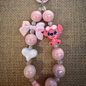 May include: A pink beaded keychain featuring a cartoon character, a bow, and a white heart. The keychain has iridescent pink beads, clear beads, and a pink clasp. The cartoon character is wearing goggles and has blue eyes.