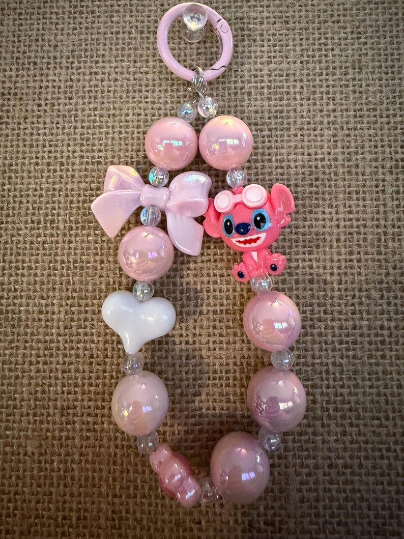 May include: A pink beaded keychain featuring a cartoon character, a bow, and a white heart. The keychain has iridescent pink beads, clear beads, and a pink clasp. The cartoon character is wearing goggles and has blue eyes.