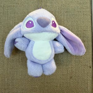May include: A plush toy of a cartoon character in shades of lavender and white, with large ears and purple eyes. The toy has a white belly and is hanging from a silver chain. The background is a textured, tan surface.