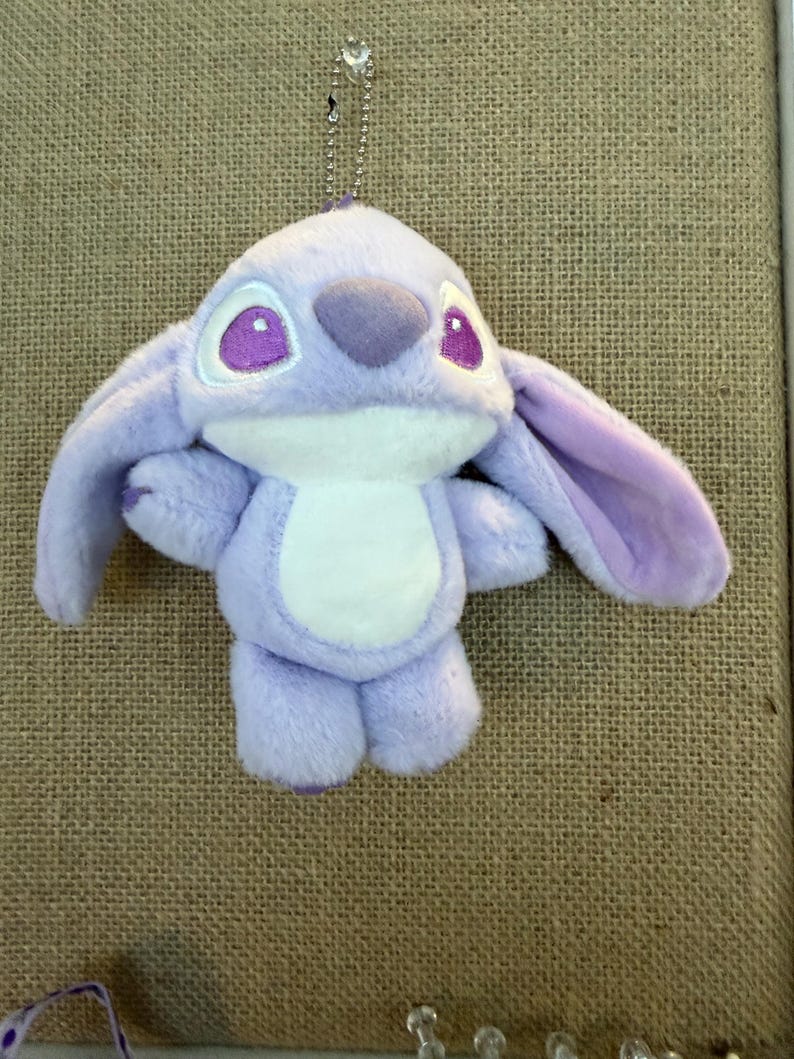 May include: A plush toy of a cartoon character in shades of lavender and white, with large ears and purple eyes. The toy has a white belly and is hanging from a silver chain. The background is a textured, tan surface.