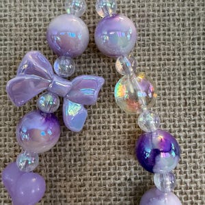 May include: A pair of dangling earrings featuring iridescent purple and clear beads. The earrings have a bow-shaped charm and round beads in varying sizes. The earrings are on a neutral background.