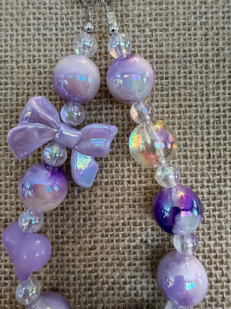 May include: A pair of dangling earrings featuring iridescent purple and clear beads. The earrings have a bow-shaped charm and round beads in varying sizes. The earrings are on a neutral background.