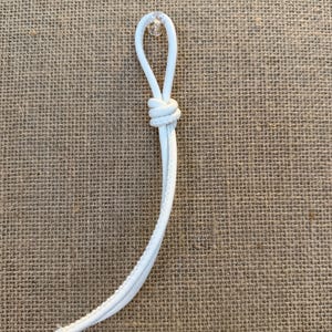 May include: A white cord with a loop and knot, possibly for a drawstring or similar application. The cord is against a textured, tan background.