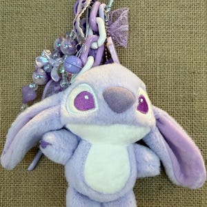 May include: A plush, lavender-colored Angel keychain with a silver clasp. The keychain features a soft, stuffed Angel character with large purple eyes, a white belly, and long ears. Decorative beads and ribbons in shades of purple and white complete the design.