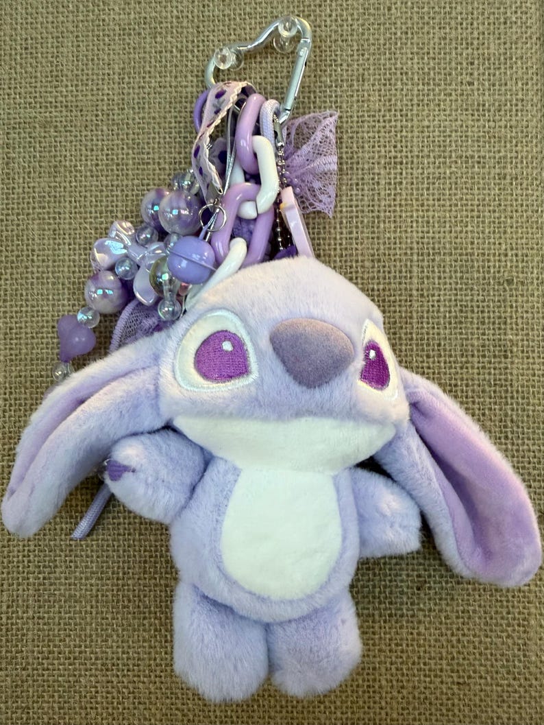 May include: A plush, lavender-colored Angel keychain with a silver clasp. The keychain features a soft, stuffed Angel character with large purple eyes, a white belly, and long ears. Decorative beads and ribbons in shades of purple and white complete the design.