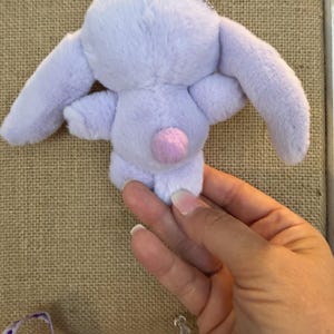 May include: A small, lavender plush bunny with long ears and a pink pom-pom tail. The bunny is attached to a silver keychain. The plush toy is held by a person's hand.