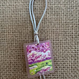 May include: A small, rectangular pendant with a landscape painting in shades of pink, purple, and green. The pendant is attached to a silver cord and a clear push pin. The artwork depicts a field and sky with a textured, impressionistic style.