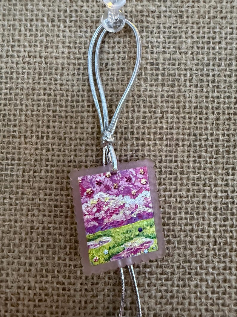 May include: A small, rectangular pendant with a landscape painting in shades of pink, purple, and green. The pendant is attached to a silver cord and a clear push pin. The artwork depicts a field and sky with a textured, impressionistic style.