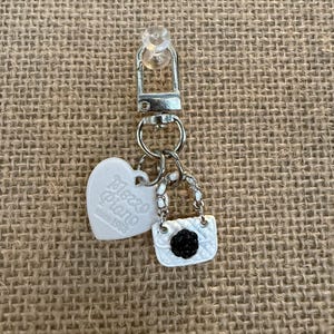 May include: A silver keychain with a small white purse charm with a black flower, a white heart charm with "Mezzo Piano" text, and a silver clasp. The keychain is on a tan burlap background.