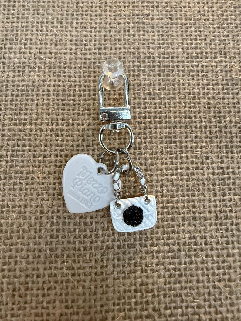 May include: A silver keychain with a small white purse charm with a black flower, a white heart charm with "Mezzo Piano" text, and a silver clasp. The keychain is on a tan burlap background.