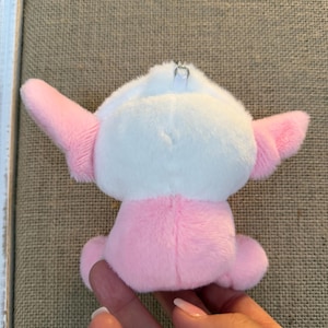 May include: A small, plush toy in the shape of a cartoon character. The toy is primarily pink and white, with pink ears and a pink body. It has a small silver metal loop attached to the top for hanging. The toy is soft and cuddly.