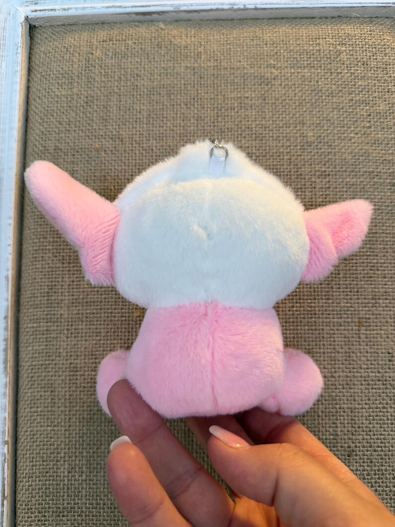 May include: A small, plush toy in the shape of a cartoon character. The toy is primarily pink and white, with pink ears and a pink body. It has a small silver metal loop attached to the top for hanging. The toy is soft and cuddly.