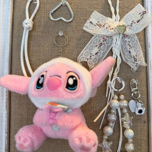 May include: A pink plush toy with large blue eyes and a white hood, surrounded by various accessories. Includes a heart-shaped carabiner, a ring, a decorative bow with floral accents, and beaded keychains. The items are displayed on a burlap background within a white framed display.