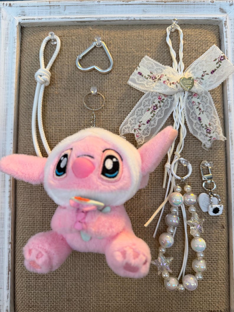 May include: A pink plush toy with large blue eyes and a white hood, surrounded by various accessories. Includes a heart-shaped carabiner, a ring, a decorative bow with floral accents, and beaded keychains. The items are displayed on a burlap background within a white framed display.