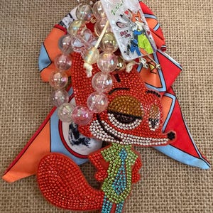 May include: A decorative keychain featuring a rhinestone-encrusted fox character wearing a green shirt and blue tie. It includes a red and blue patterned fabric bow, a strand of iridescent beads, and a rectangular charm with Zootopia text.