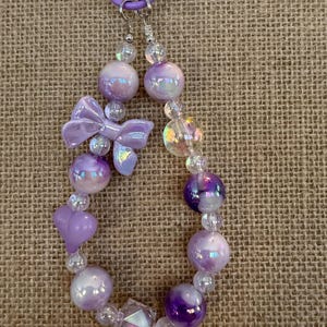 May include: A beaded keychain featuring iridescent purple beads, a bow, and heart-shaped beads. The keychain is attached to a purple ring. The beads vary in size and shape, creating a unique and eye-catching accessory.