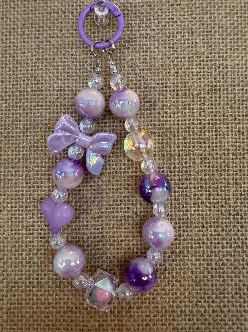 May include: A beaded keychain featuring iridescent purple beads, a bow, and heart-shaped beads. The keychain is attached to a purple ring. The beads vary in size and shape, creating a unique and eye-catching accessory.