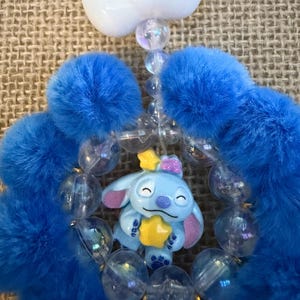 May include: A blue and white beaded charm featuring a cartoon character holding a yellow star. The charm has blue fluffy accents and a white bow. Clear beads and a white bow complete the design.