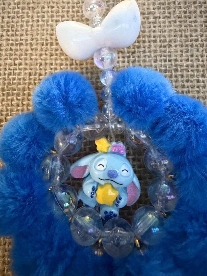 May include: A blue and white beaded charm featuring a cartoon character holding a yellow star. The charm has blue fluffy accents and a white bow. Clear beads and a white bow complete the design.