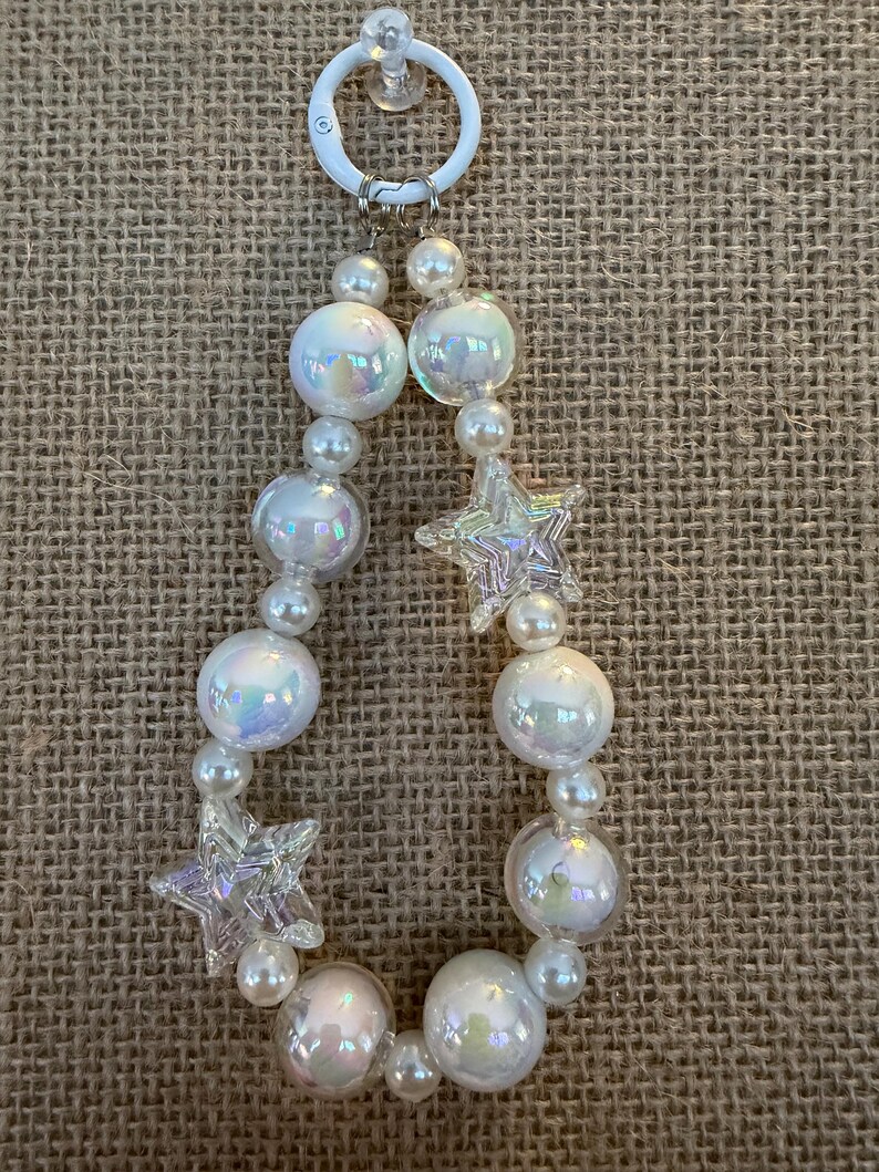 May include: A beaded keychain featuring iridescent, round beads and smaller pearl-like beads. Two clear, star-shaped charms are interspersed among the beads. The keychain has a white ring and suction cup for attachment.
