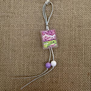 May include: A small, square pendant with a landscape painting of purple flowers and green grass. The pendant is attached to a silver cord with two beads, one purple and one white. The background is a burlap texture.