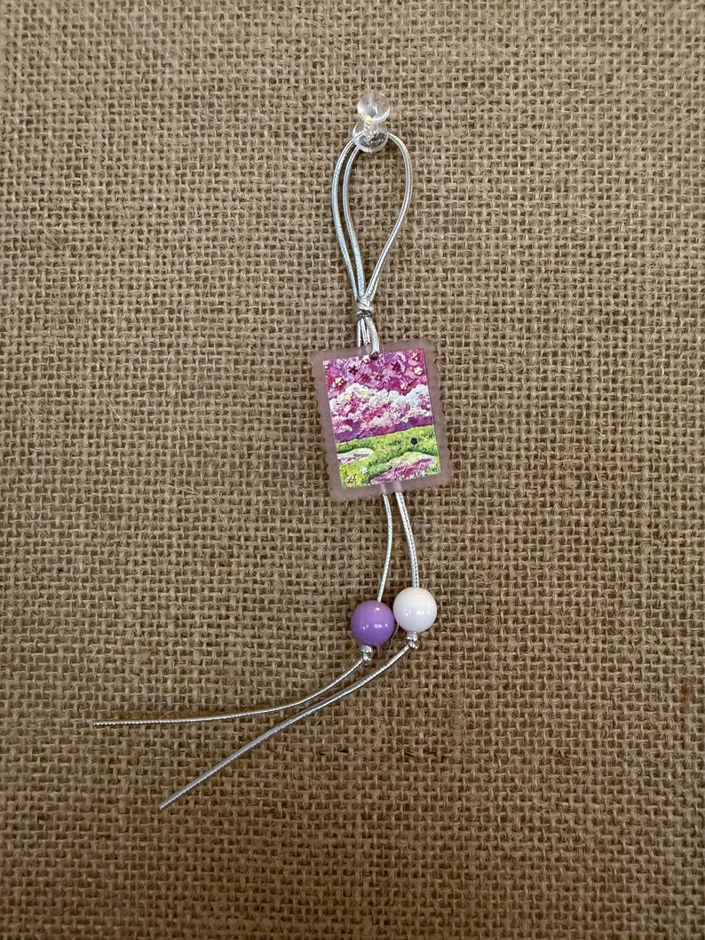 May include: A small, square pendant with a landscape painting of purple flowers and green grass. The pendant is attached to a silver cord with two beads, one purple and one white. The background is a burlap texture.