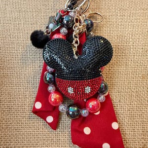 May include: A keychain featuring a black and red rhinestone-encrusted mouse head charm, a red fabric ribbon with white polka dots, and a collection of black, red, and clear beads. The assembly is attached to a gold-toned star-shaped metal clasp.