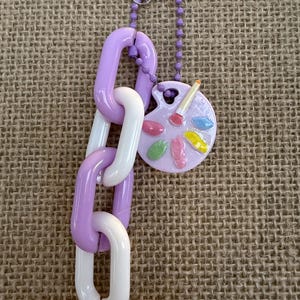 May include: A decorative keychain featuring a chain of purple and white links, connected to a small, round palette charm. The palette has colorful paint splotches and a paintbrush. The keychain is attached to a clear suction cup.