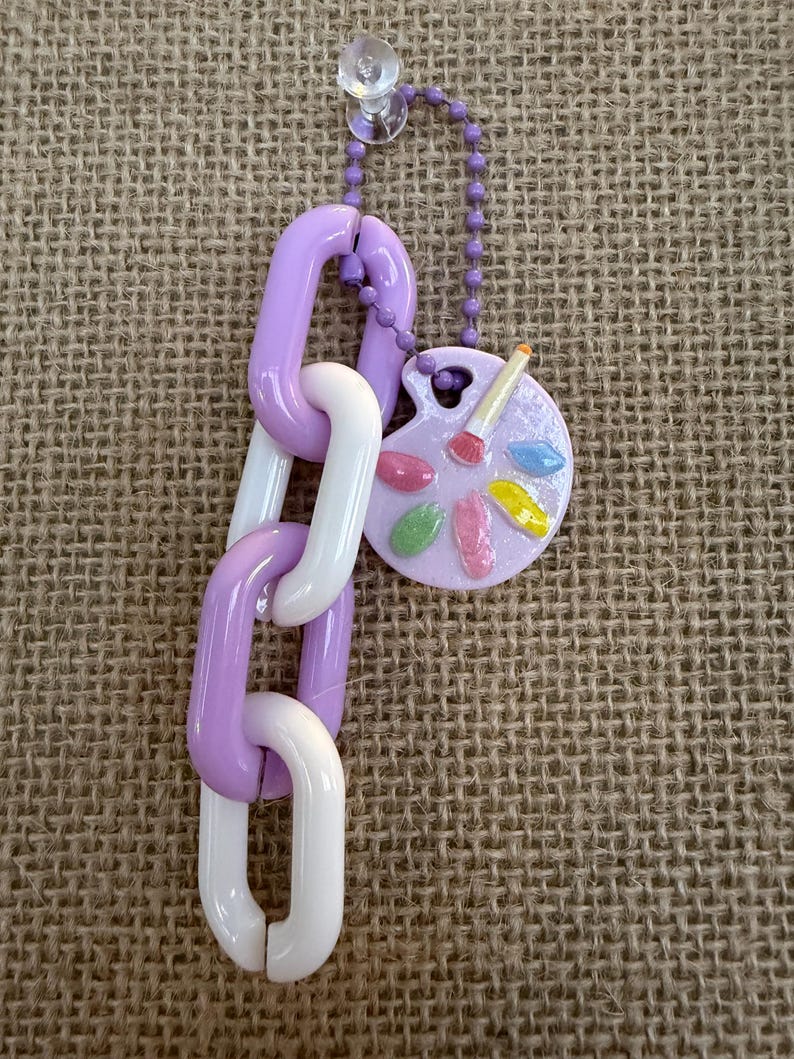 May include: A decorative keychain featuring a chain of purple and white links, connected to a small, round palette charm. The palette has colorful paint splotches and a paintbrush. The keychain is attached to a clear suction cup.