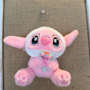 May include: A pink plush toy with large black eyes, a pink nose, and large ears. The toy has a white hood and is holding a flower. The toy is attached to a silver keychain.