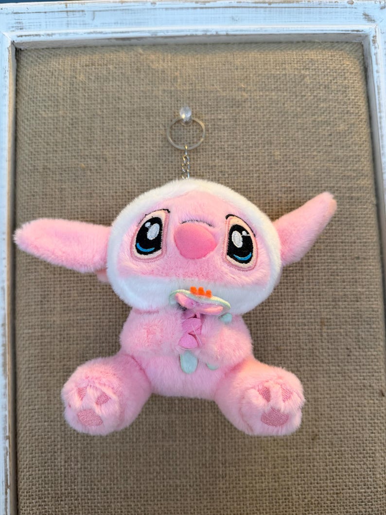 May include: A pink plush toy with large black eyes, a pink nose, and large ears. The toy has a white hood and is holding a flower. The toy is attached to a silver keychain.