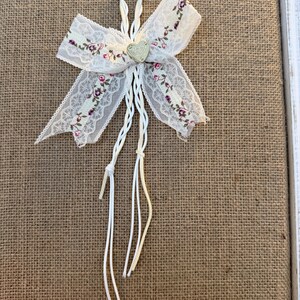 May include: A decorative white lace bow with a floral pattern in pink and purple. The bow has a small heart-shaped charm in the center and white string accents. The bow is attached to a burlap background.