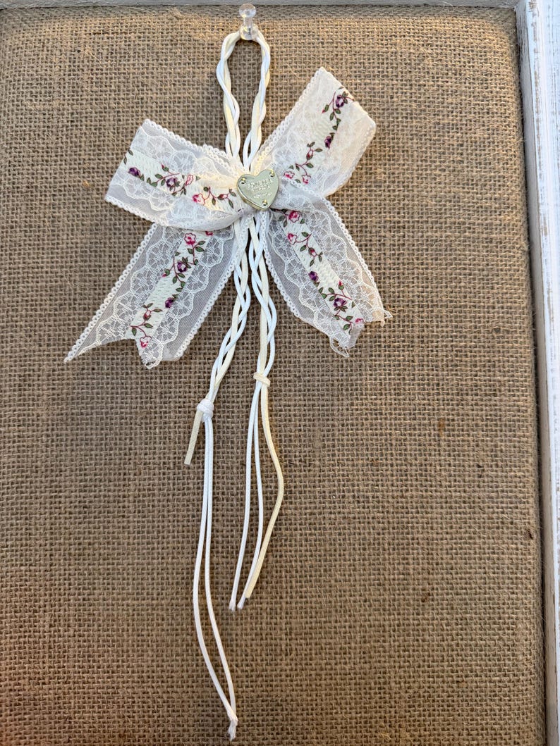 May include: A decorative white lace bow with a floral pattern in pink and purple. The bow has a small heart-shaped charm in the center and white string accents. The bow is attached to a burlap background.