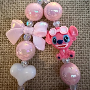 May include: A pink beaded necklace featuring a cartoon character, bows, and heart-shaped beads. The necklace has iridescent pink and clear beads, with a pink bow and a white heart. The cartoon character is pink with blue eyes and goggles.