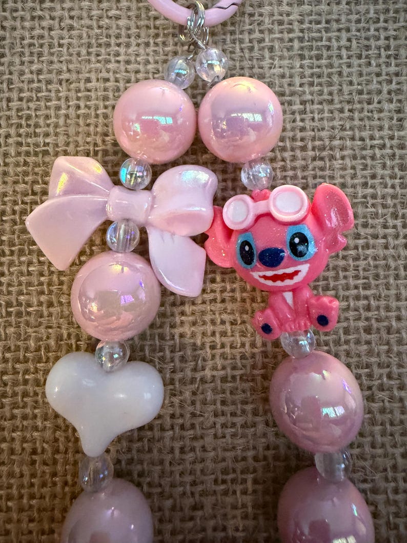 May include: A pink beaded necklace featuring a cartoon character, bows, and heart-shaped beads. The necklace has iridescent pink and clear beads, with a pink bow and a white heart. The cartoon character is pink with blue eyes and goggles.