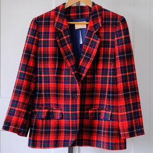 May include: A red, blue, and green tartan blazer with a single-button fastening and two front pockets. The blazer is suspended on a wooden hanger. The interior lining is a solid navy blue. The background is white.