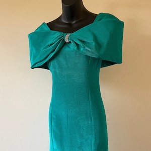 Puede incluir: Vestido turquesa con hombros descubiertos y un gran lazo en el escote, adornado con pedrer&iacute;a. El vestido tiene una silueta ajustada y un largo hasta la rodilla, ideal para eventos formales. El vestido mide unos 100 cm de largo.