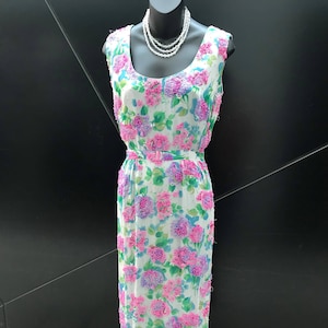 Peut inclure: Robe longue sans manches avec un motif floral de fleurs roses et violettes sur fond blanc. La robe est dotée d'une ceinture assortie et est présentée sur un mannequin avec un collier de perles.