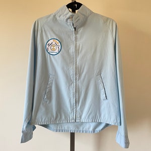 Puede incluir: Chaqueta azul claro con cierre de cremallera completo, dos bolsillos delanteros y cuello alzado. Un parche circular en el pecho dice "Blue Mountain Boys" con texto adicional. La chaqueta está colgada de una percha negra.