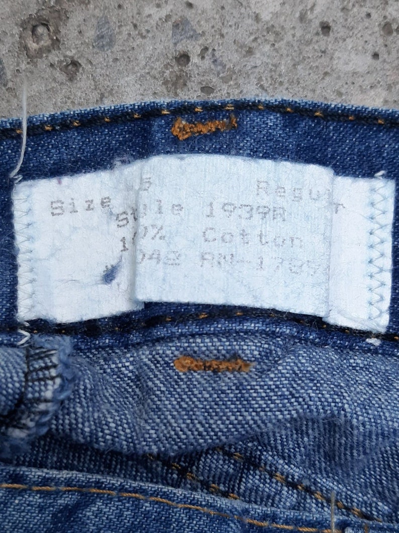 Puede incluir: Primer plano de unos vaqueros azules con una etiqueta blanca. La etiqueta dice "Size 5 Regular", "Style 1939R" y "100% Cotton". Los vaqueros tienen costuras visibles y un bot&oacute;n.