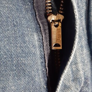 May include: Close-up of a denim garment with a partially open zipper. The zipper is metallic with the word "GRIPPER ZIPPER" embossed on the pull tab. The denim fabric is a light blue wash.