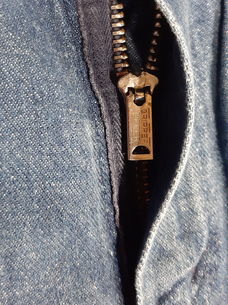 May include: Close-up of a denim garment with a partially open zipper. The zipper is metallic with the word "GRIPPER ZIPPER" embossed on the pull tab. The denim fabric is a light blue wash.