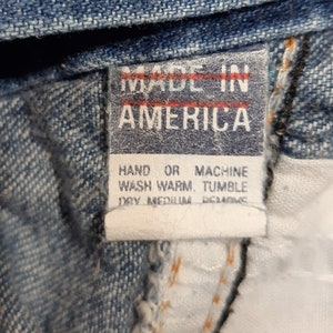 Puede incluir: Primer plano de una etiqueta de prenda vaquera. La etiqueta dice "MADE IN AMERICA" en letras may&uacute;sculas blancas sobre un fondo rojo. Las instrucciones de lavado est&aacute;n impresas debajo en texto negro.