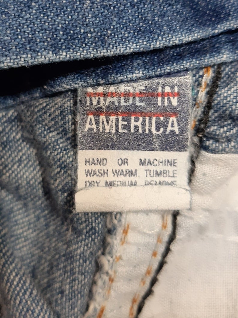 Puede incluir: Primer plano de una etiqueta de prenda vaquera. La etiqueta dice "MADE IN AMERICA" en letras may&uacute;sculas blancas sobre un fondo rojo. Las instrucciones de lavado est&aacute;n impresas debajo en texto negro.