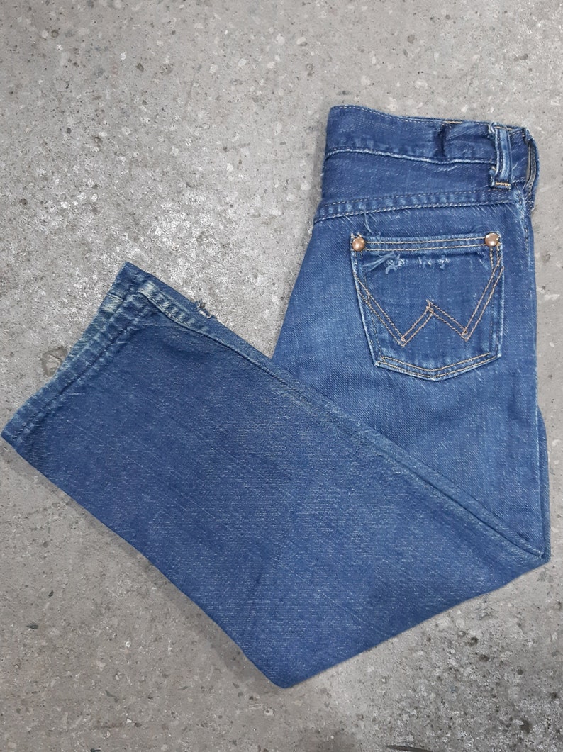 May include: Blue denim jeans with a classic five-pocket design. Features a distressed pocket and the Wrangler logo on the back pocket. The jeans are folded and laid flat on a gray surface.