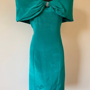 Puede incluir: Vestido turquesa con hombros descubiertos y un gran lazo en el escote, adornado con pedrer&iacute;a. El vestido tiene una silueta ajustada y un dobladillo a la rodilla. Se muestra en un maniqu&iacute; negro. El vestido es de color turquesa.