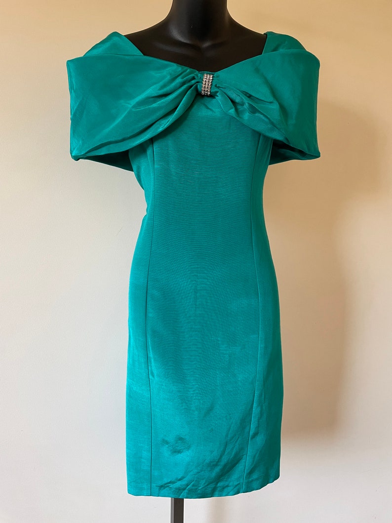 Puede incluir: Vestido turquesa con hombros descubiertos y un gran lazo en el escote, adornado con pedrer&iacute;a. El vestido tiene una silueta ajustada y un dobladillo a la rodilla. Se muestra en un maniqu&iacute; negro. El vestido es de color turquesa.