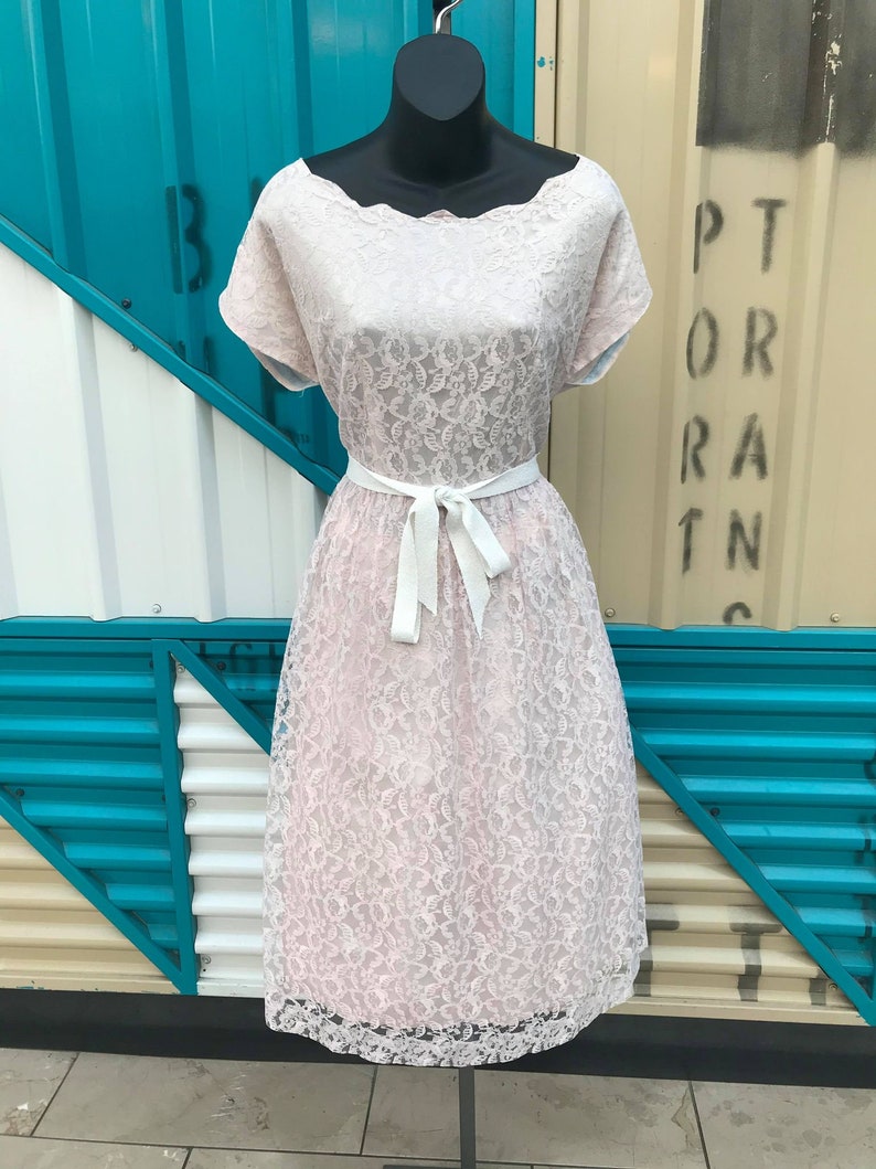 Puede incluir: Vestido de encaje rosa claro sobre un maniqu&iacute;. El vestido presenta un escote festoneado, mangas cortas y una falda a la rodilla. Un cintur&oacute;n de cinta blanca ci&ntilde;e la cintura. El vestido se exhibe sobre un fondo de paneles turquesa y crema.